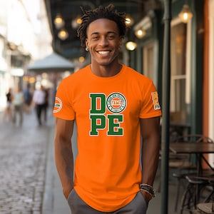 May include: Bright orange t-shirt with the word "DOPE" in green block letters. The letter "O" is replaced with a circular emblem. Additional logos are on the left sleeve and right shoulder. The shirt is worn by a person.