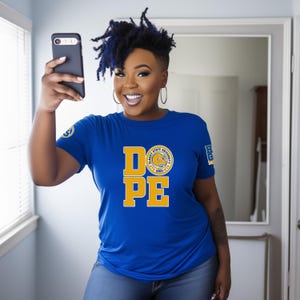 Könnte beinhalten: Blaues T-Shirt mit dem Wort "DOPE" in gelben Blockbuchstaben und einem kreisförmigen Logo. Das Shirt hat auch ein Logo auf dem linken Ärmel. Die Person hält ein schwarzes Smartphone.