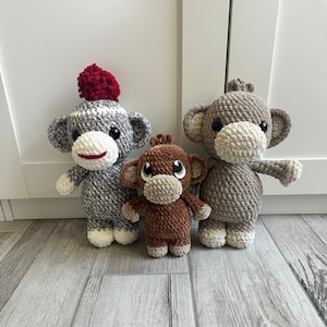 Könnte beinhalten: Drei gehäkelte Affenspielzeuge. Ein Affe ist grau mit einer roten Bommelmütze, ein anderer ist braun und der dritte ist grau mit einem weißen Gesicht.