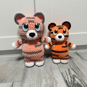 May include: Two crocheted tiger plush toys. One is peach and grey, the other is orange and black. Both have large, cartoonish eyes, white paws, and are standing upright. The larger tiger has grey ears, while the smaller one has black ears.