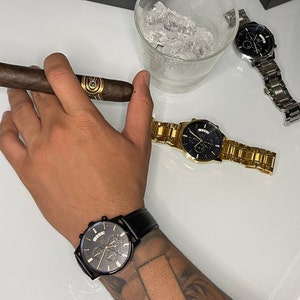 Pode incluir: Uma variedade de rel&oacute;gios de luxo em tons de preto e dourado, e um rel&oacute;gio prateado. Um charuto aceso &eacute; segurado na m&atilde;o. Um copo de gelo est&aacute; no fundo. Os rel&oacute;gios t&ecirc;m v&aacute;rios estilos de pulseira.