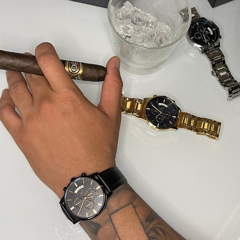 Pode incluir: Uma variedade de rel&oacute;gios de luxo em tons de preto e dourado, e um rel&oacute;gio prateado. Um charuto aceso &eacute; segurado na m&atilde;o. Um copo de gelo est&aacute; no fundo. Os rel&oacute;gios t&ecirc;m v&aacute;rios estilos de pulseira.