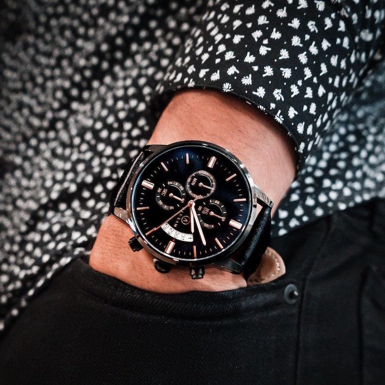 Pode incluir: Um rel&oacute;gio de pulso preto com mostrador azul escuro e detalhes em ouro rosa. O rel&oacute;gio tem uma pulseira de couro preta e uma caixa de a&ccedil;o inoxid&aacute;vel. O rel&oacute;gio est&aacute; em um pulso, com uma manga de camisa estampada em preto e branco ao fundo.