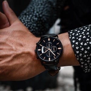 Pode incluir: Rel&oacute;gio de pulso preto com uma pulseira de couro preta e um mostrador escuro com detalhes dourados. O rel&oacute;gio tem v&aacute;rios mostradores e um visor de data. O rel&oacute;gio est&aacute; em um pulso, com uma manga de camisa estampada no fundo.