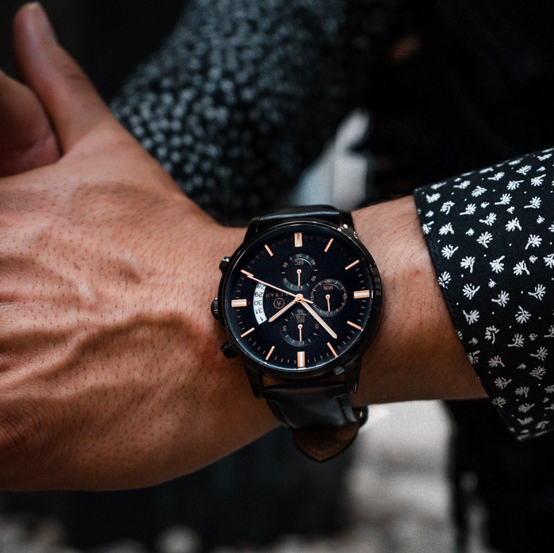 Pode incluir: Rel&oacute;gio de pulso preto com uma pulseira de couro preta e um mostrador escuro com detalhes dourados. O rel&oacute;gio tem v&aacute;rios mostradores e um visor de data. O rel&oacute;gio est&aacute; em um pulso, com uma manga de camisa estampada no fundo.