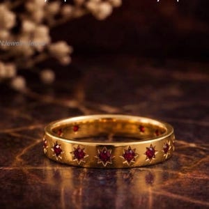 May include: A gold band ring featuring a series of star-shaped settings, each containing a small, round, deep red ruby. The metal surface is polished and smooth, with the text "GemsNJewelryJaipur" visible in the background of the image.