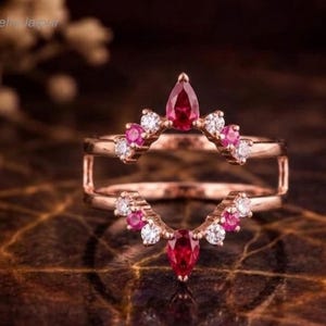 May include: A rose gold ring guard featuring two symmetrical bands connected at the sides. Each band is adorned with a central pear-cut red gemstone flanked by alternating round-cut red and clear white stones in a chevron-inspired arrangement.