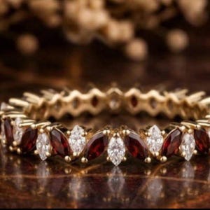 May include: A gold ring with alternating marquise-cut red and white gemstones. The ring's band is a continuous loop, showcasing the gemstones in a repeating pattern. The ring is set on a dark, reflective surface.