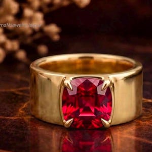 Peut inclure: Une large bague en or dotée d'une grande pierre précieuse rouge foncé taille coussin, fixée au centre par quatre griffes. La bague présente une finition métallique lisse et polie ainsi qu'un profil épais et substantiel.