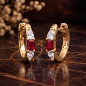 Pode incluir: Um par de brincos de argola dourados com carimbo 14K. Cada brinco apresenta uma pedra vermelha retangular central, ladeada por duas pedras brancas em forma de pêra. Fundo desfocado.
