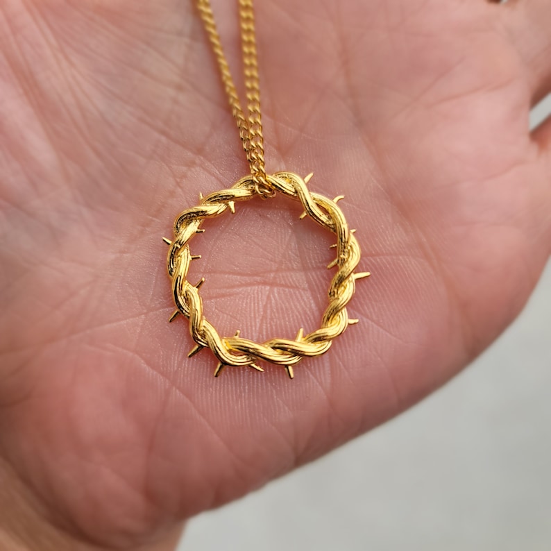 May include: A gold-toned necklace with a crown of thorns pendant. The pendant is a circular design with a twisted rope-like texture and sharp points.