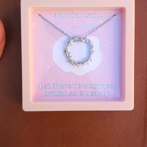 May include: A pink box with a silver necklace inside. The necklace has a circular pendant with a barbed wire design. The box has the text "And God said... Let there be elegance, brilliance & beauty." printed on it.