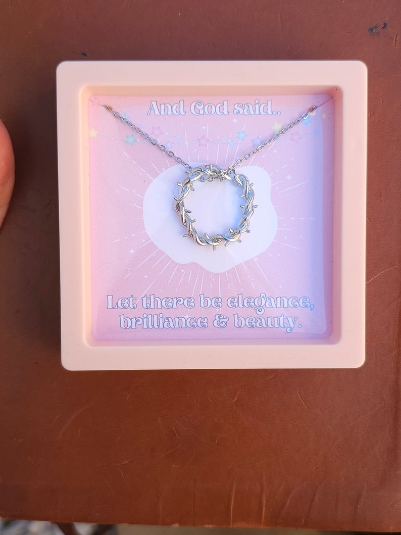May include: A pink box with a silver necklace inside. The necklace has a circular pendant with a barbed wire design. The box has the text "And God said... Let there be elegance, brilliance & beauty." printed on it.