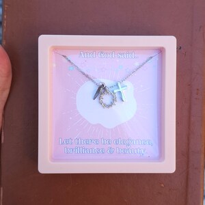 May include: A pink box with a silver necklace inside. The necklace has a cross, a crown of thorns, and a quote that reads "And God said... Let there be elegance, brilliance & beauty."