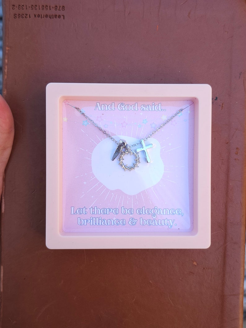May include: A pink box with a silver necklace inside. The necklace has a cross, a crown of thorns, and a quote that reads "And God said... Let there be elegance, brilliance & beauty."