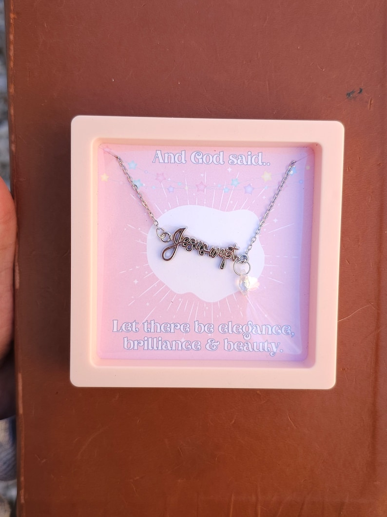 May include: A pink box with a silver necklace inside. The necklace has a charm that says "Jesus wept" and a small pearl charm. The box has the text "And God said... Let there be elegance, brilliance & beauty."