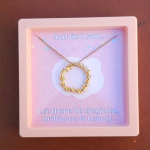 May include: A pink box with a gold necklace inside. The necklace has a circular pendant with a barbed wire design. The box has a pink background with white stars and the text "And God said... Let there be elegance, brilliance & beauty."