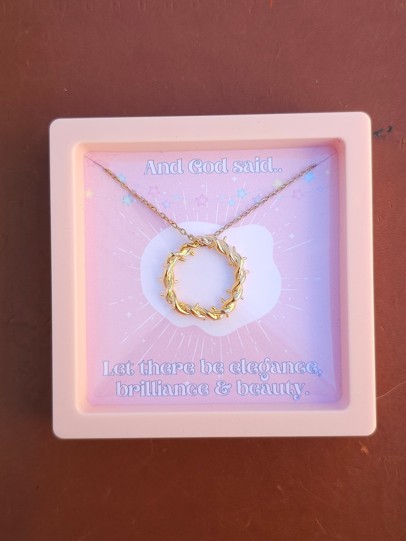 May include: A pink box with a gold necklace inside. The necklace has a circular pendant with a barbed wire design. The box has a pink background with white stars and the text "And God said... Let there be elegance, brilliance & beauty."