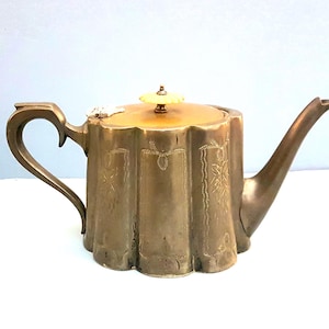 Op de afbeelding: Antieke messing theepot met een hartvormige handgreep en een gebogen tuit. De theepot heeft een geschulpte rand en decoratieve bloemetsingen. Het deksel is bekroond met een crèmekleurige knop. Een vintage theeaccessoire.