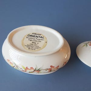 Puede incluir: Un recipiente ovalado de cerámica blanca para polvos, con tapa. El recipiente y la tapa están decorados con motivos florales en tonos naranja, rosa y verde. El recipiente tiene una etiqueta que dice "ORIENTAL Dusting Powder" y pesa 90g.
