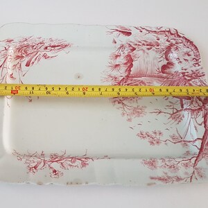 Peut inclure: Plat rectangulaire en céramique blanche avec un motif floral et paysager rouge. Le plat mesure environ 30 cm de long. Les bords ont un design festonné.