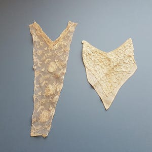 May include: Two pieces of delicate, off-white lace fabric. One piece is a long, narrow strip with a V-neck shape and floral embroidery. The other piece is a triangular shape with a more textured lace pattern.