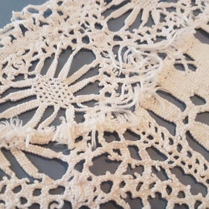 May include: Close-up of a white, handmade crochet doily with a floral design. The doily features a detailed openwork pattern with a fringed edge. The intricate design is set against a muted blue background, highlighting the delicate texture.