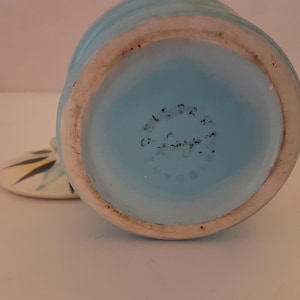 May include: A vintage ceramic jar in light blue with a matching lid. The jar's base is a lighter shade of blue, with a white rim. The lid has a floral design in yellow, black, and white. The jar's bottom has text.