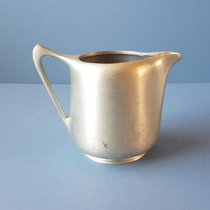 Puede incluir: Una pequeña jarra de crema de color plateado con un asa curva y un pico. La jarra tiene un cuerpo redondeado y se asienta sobre una pequeña base. La jarra de crema es de metal y tiene un diseño simple y elegante.