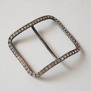 May include: A vintage silver-toned buckle with a square, slightly curved shape. The buckle is encrusted with small, clear rhinestones around the outer edge. A single bar runs across the center of the opening.