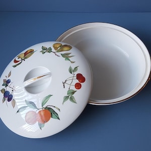 Puede incluir: Plato de servir de cerámica blanca con tapa. La tapa tiene un borde dorado y un diseño floral con frutas, incluyendo cerezas, peras y naranjas. El plato está vacío.