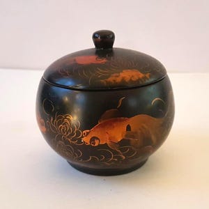 Op de afbeelding: Een zwarte, ronde container met deksel en een donkerbruine knop. De container is versierd met oranje en gouden koi-vissen en bloemmotieven. De container is waarschijnlijk gemaakt van hout of een vergelijkbaar materiaal.