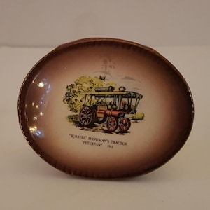 Puede incluir: Un plato decorativo ovalado marrón con una ilustración colorida de un tractor de vapor antiguo. El plato tiene un borde marrón oscuro y el texto "BURRELL SHOWMAN'S TRACTOR PETERPAN 1912".