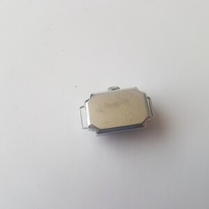 以下が含まれることがあります： シルバーカラーのケースと明るい色の文字盤を備えた、ヴィンテージの長方形の腕時計。 時計には、小さなテクスチャード加工のリュウズと、バンドを取り付けるための2つのラグが付いています。 時計は白い背景に配置されています。