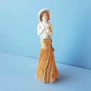 以下が含まれることがあります： わらのほうきスカートを着たヴィンテージ磁器人形。人形は白い帽子と緑のドットの白いブラウスを着ています。スカートは金色のわらでできています。