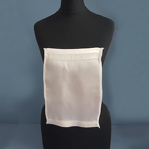 Pode incluir: Um lenço de linho branco retangular com bordas bordadas e detalhes de costura. O lenço é exibido em um manequim preto contra um fundo azul, mostrando suas linhas limpas e seu design clássico.