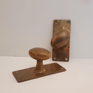 Pode incluir: Duas maçanetas e placas traseiras de porta de bronze antigo. Um botão é montado em uma placa traseira retangular, o outro é separado. O hardware tem uma pátina envelhecida, com botões ovais e placas traseiras retangulares. A ferragem da porta é provavelmente do início do século 20.