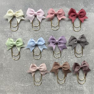 Könnte beinhalten: Eine Auswahl an farbenfrohen Schleifen aus Band, die an goldfarbenen Büroklammern befestigt sind. Die Schleifen sind in verschiedenen Pastelltönen wie Rosa, Blau, Grün und Lila gehalten. Die Büroklammern sind auf einer grauen Oberfläche angeordnet.