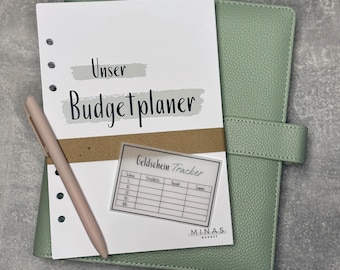 Könnte beinhalten: Ein weißer Planer mit dem Text "Unser Budgetplaner" auf dem Cover. Eine transparente Plastikfolie mit dem Text "Geldschein Tracker" und einer Tabelle ist auf dem Cover befestigt. Der Planer ist geschlossen und hat einen grünen Lederumschlag.