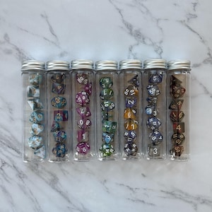 Könnte beinhalten: Sieben transparente Glasröhren mit silbernen Deckeln, jede gefüllt mit einem Satz polyedrischer Würfel. Die Würfel sind in verschiedenen Farben, darunter Blau, Lila, Grün und Braun, mit weißen oder goldenen Zahlen. Würfel für Tischspiele.