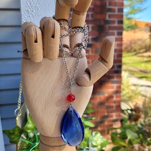 May include: A silver chain necklace with a teardrop-shaped blue crystal pendant and a small red bead.