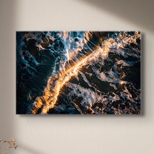 Puede incluir: Una vista aérea de las olas del océano rompiendo contra la costa, creando un patrón de remolino de espuma blanca y agua azul oscuro. La luz del sol se refleja en el agua, creando un brillo dorado.