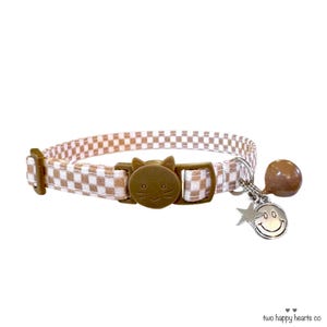 May include: A cat collar featuring a brown and white gingham pattern. The collar has a brown cat-shaped buckle and a silver charm with a star and a smiling face. A brown bell is attached.
