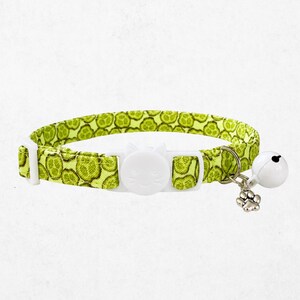 Puede incluir: Un collar para mascota de color verde lima con un patrón repetitivo de rodajas de pepino. Incluye una hebilla de plástico blanco con forma de cara de gato, un dije de metal plateado con forma de huella y un pequeño cascabel de color blanco.
