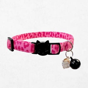 A pink leopard print cat collar with a black buckle and bell attached.