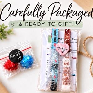 a carefully packaged gift with a note that says "Carefully Packaged & READY TO GIFT!" The gift includes a small bag of red and blue pom poms, a package of what appears to be a watch, and a note that says "Two Heart".