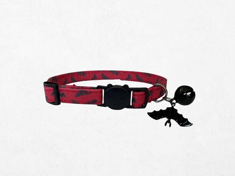 A red cat collar with a bell and a bat-themed charm is shown against a white background.