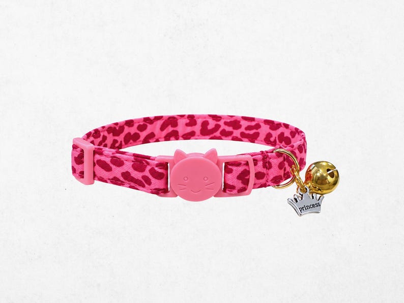 A pink cat collar with a bell and a charm featuring a cat's face.