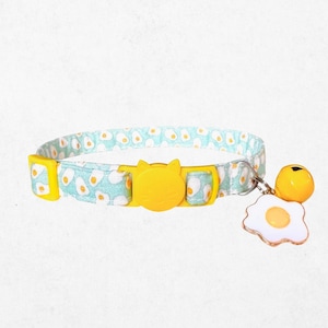 May include: A cat collar with a light blue and white avocado pattern. The yellow buckle is shaped like a cat's face. A yellow bell and a fried egg charm dangle from the collar.
