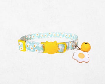 May include: A cat collar with a light blue and white avocado pattern. The yellow buckle is shaped like a cat's face. A yellow bell and a fried egg charm dangle from the collar.